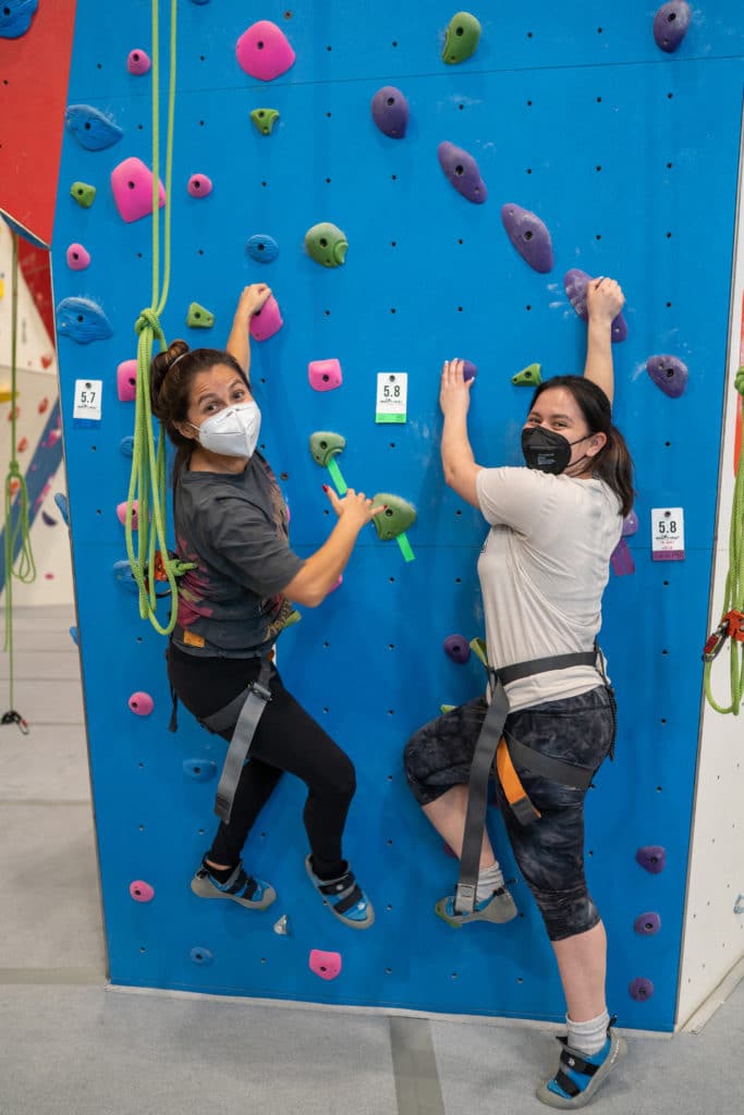 Two people face the camera before starting their climb at a climbing gym.
