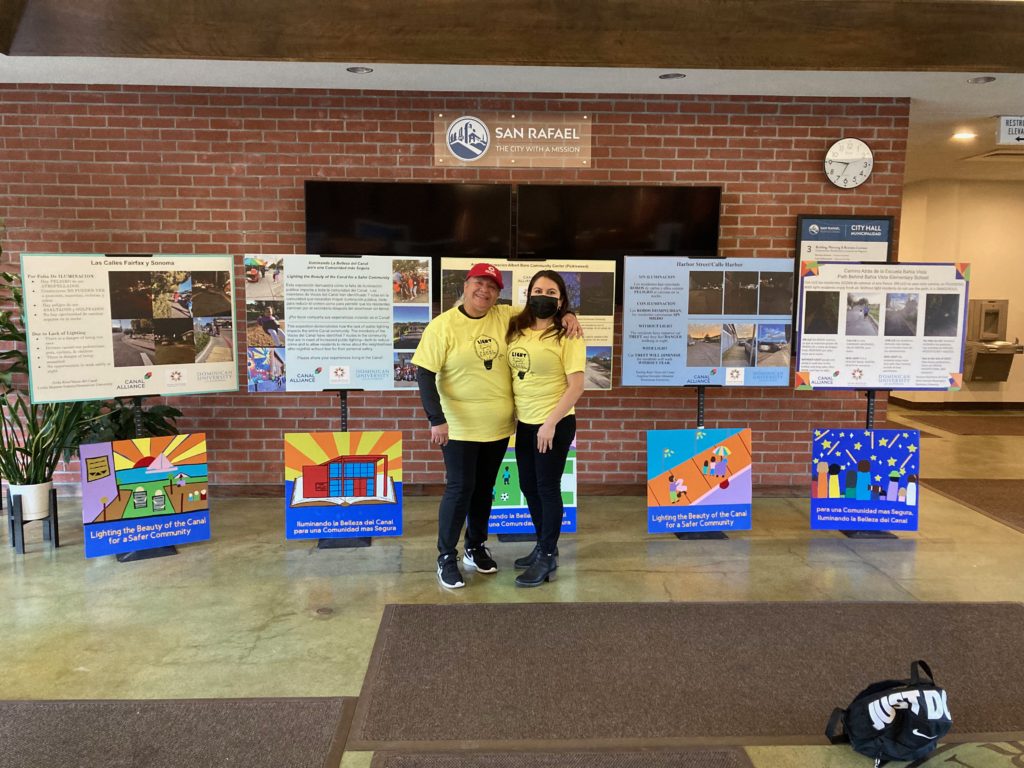 Two people in yellow shirts standing in the lobby of San Rafael City Hall in front of presentation materials.